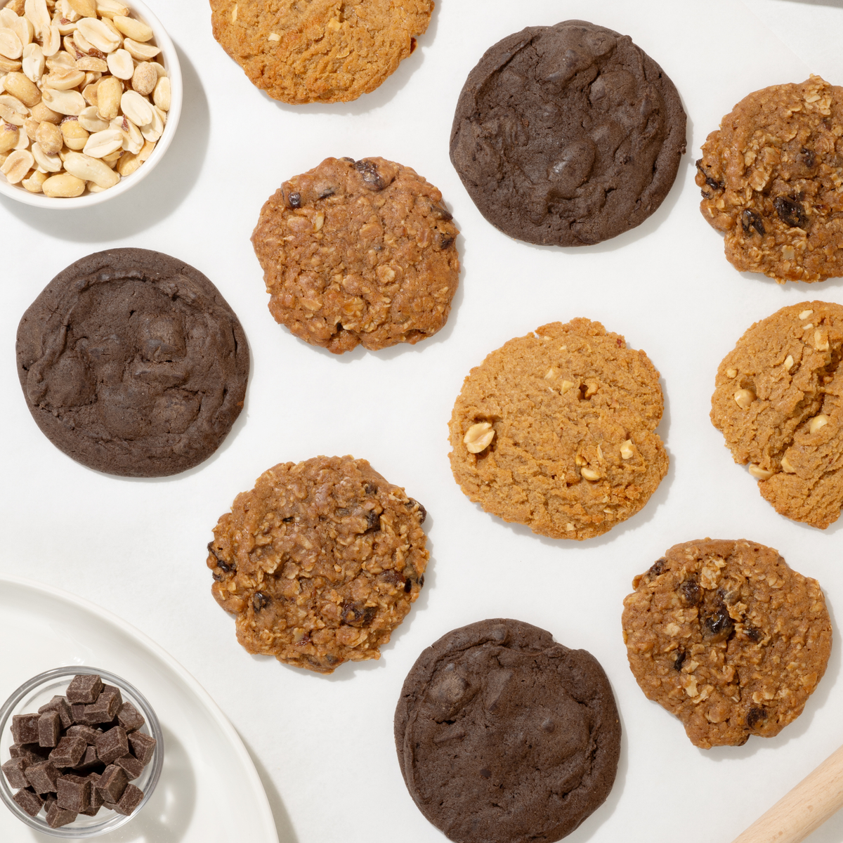 An overhead flat-lay of assorted round Fields Good cookies on a white surface, including chocolate chip, oatmeal, and peanut butter cookies, with small bowls of peanuts and chocolate chunks nearby.