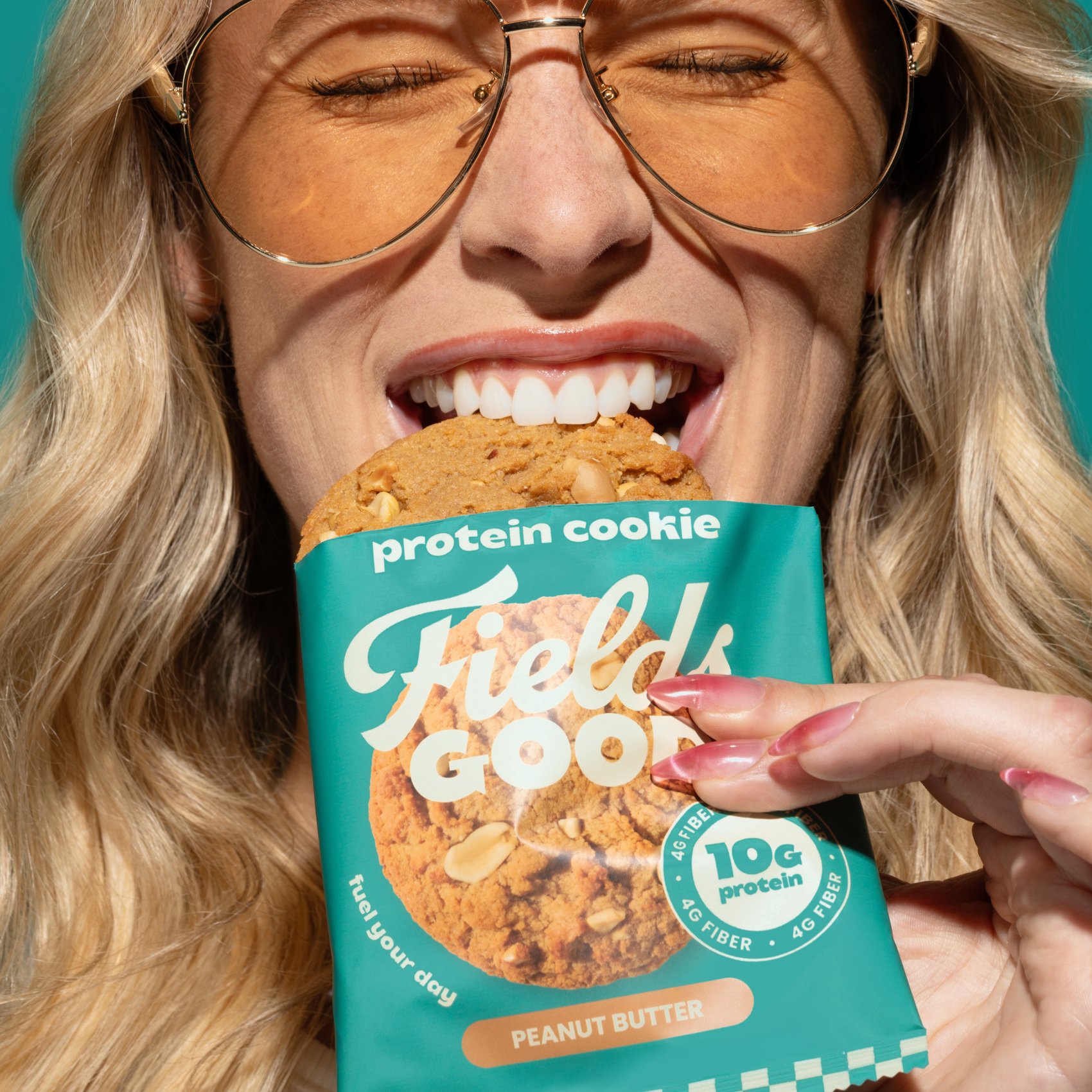 Person holding a Fields Good protein cookie package with a cookie inside, against a teal background