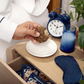 Person holding a cookie next to a plate with a blue alarm clock, glass, and eye mask on a wooden surface.