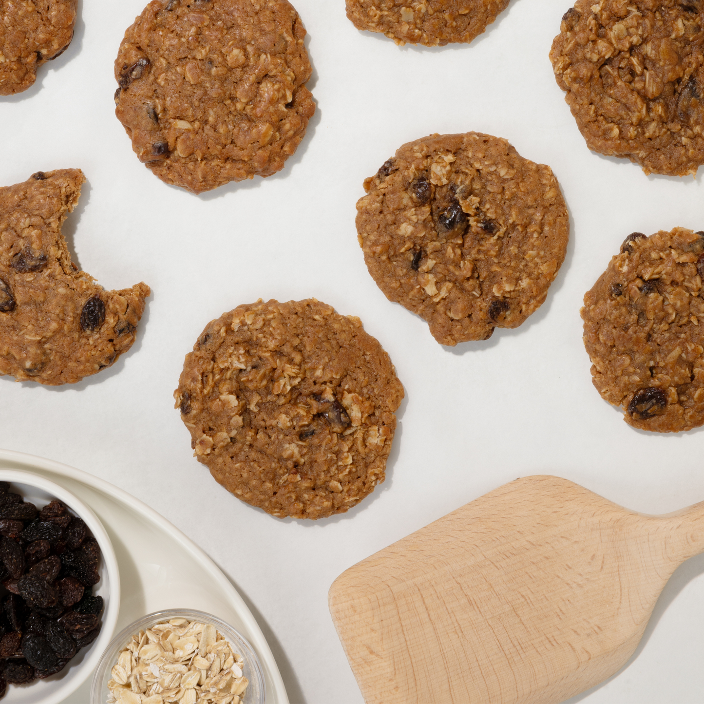 Oatmeal raisin cookies on a white surface with a wooden spatula and ingredients.