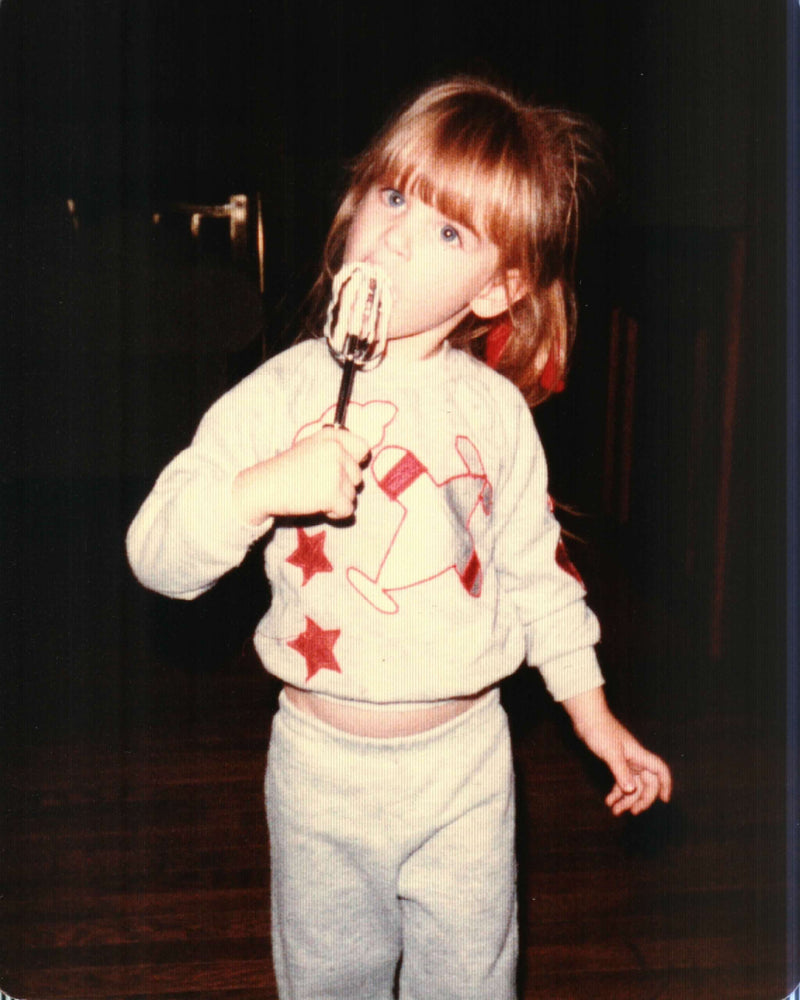 Vintage childhood photo of a young girl in a star-print sweatshirt licking cake batter off a mixer beater.